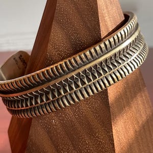 K&ouml;nnte beinhalten: Ein breites, silberfarbenes Armreif mit geometrischem Muster. Das Armband ist aus Metall und hat eine strukturierte Oberfl&auml;che.
