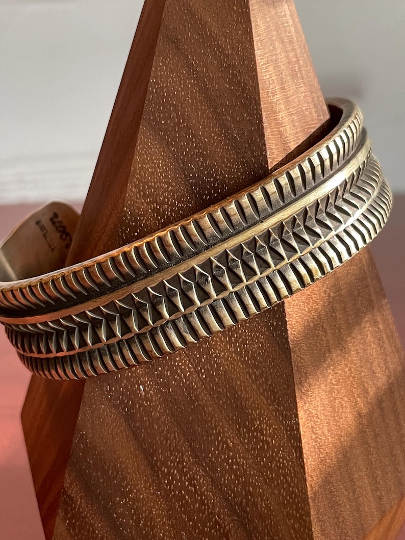K&ouml;nnte beinhalten: Ein breites, silberfarbenes Armreif mit geometrischem Muster. Das Armband ist aus Metall und hat eine strukturierte Oberfl&auml;che.
