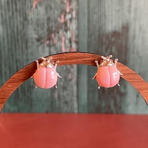 May include: A pair of pink beetle-shaped earrings with silver-colored accents. The earrings are displayed on a wooden stand against a green and brown background. The earrings have a glossy finish and are designed to resemble insects.