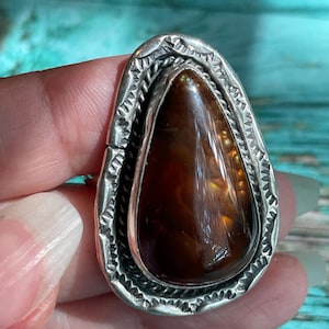 May include: A teardrop-shaped, silver-toned ring with a large, polished, brown gemstone. The gemstone has a glossy, translucent appearance with internal patterns. The ring's setting features a detailed, textured border.