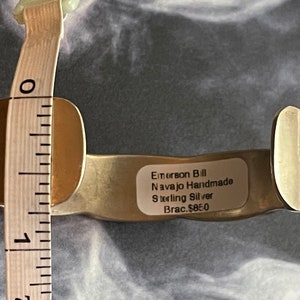 May include: A silver Navajo handmade bracelet with a label that reads "Emerson Bill Navajo Handmade Sterling Silver Brac. $85.0" and a measuring tape showing the bracelet is 2 1/2 inches in diameter.