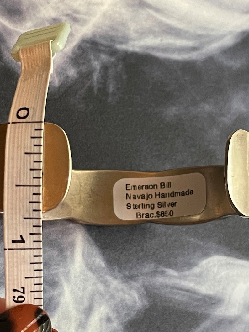 May include: A silver Navajo handmade bracelet with a label that reads "Emerson Bill Navajo Handmade Sterling Silver Brac. $85.0" and a measuring tape showing the bracelet is 2 1/2 inches in diameter.