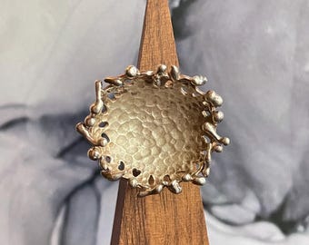 May include: A silver ring with a textured, circular design. The ring has a raised, textured center and a jagged, irregular outer edge.