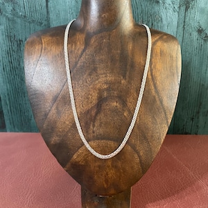 May include: A silver-toned chain necklace featuring a series of small, interlocking links that create a textured, uniform appearance. The necklace is displayed in a simple, continuous loop, showcasing the metallic finish and the consistent pattern of the chain links throughout its entire length.