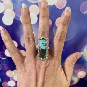May include: A silver ring featuring a rectangular turquoise and black stone. The ring has a detailed silver setting with a decorative border. The turquoise stone has a striking blue-green color, and the ring is worn on a finger.