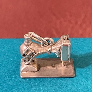 May include: A miniature silver-toned metal charm shaped like a vintage sewing machine. The charm features a rectangular base, a detailed arm with a lattice-style cutout pattern on the side, and a small jump ring attached to the top for hanging.