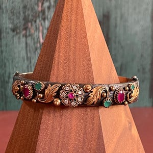 May include: Ornate silver and gold bangle bracelet with a floral and leaf design. The bracelet features a central flower with a red gemstone and surrounding diamonds, along with red and green gemstones. The bracelet is displayed on a wooden stand.