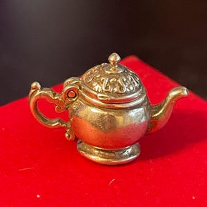 May include: A small, silver-colored teapot charm with intricate detailing. The teapot features a curved spout, a handle, and a decorative lid. It is set against a vibrant red surface, with a dark background.