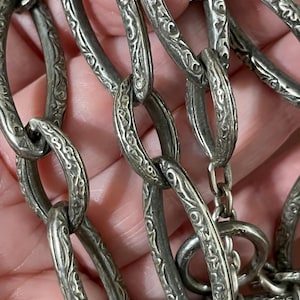 May include: A silver-toned metal chain necklace featuring elongated oval links with a detailed, raised, repeating scrollwork pattern. The necklace includes a toggle clasp mechanism with a matching textured finish on both the bar and the ring components.