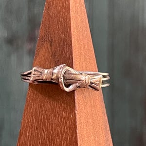 May include: A silver-toned ring featuring a bow design. The ring has a detailed knot in the center and delicate bow details on either side. The ring is displayed on a wooden stand with a natural wood grain finish.