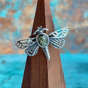 May include: A silver dragonfly ring with a green stone center, displayed on a wooden stand. The ring's wings are intricately detailed, and the stone has a speckled appearance. The background is a mix of blue and brown.