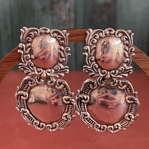 May include: A pair of silver-toned earrings. Each earring features a square top with an ornate, swirling design and a circular, reflective center. Below, a larger, similar design with a circular center completes the drop earring.