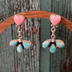 May include: A pair of silver earrings with a pink heart-shaped stone at the top and three turquoise stones at the bottom. The earrings are displayed on a wooden stand against a green background.