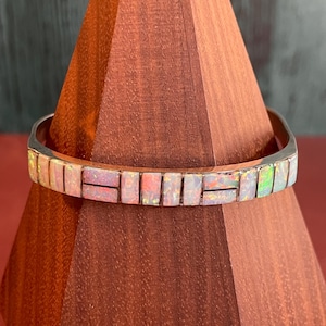 May include: A silver bracelet with a rectangular opal inlay. The opal stones are white with iridescent flashes of color. The bracelet is displayed on a wooden stand.