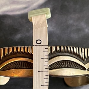 May include: A silver and brass Native American style cuff bracelet with a decorative design. The bracelet is measured with a ruler, showing it is approximately 1 inch wide.