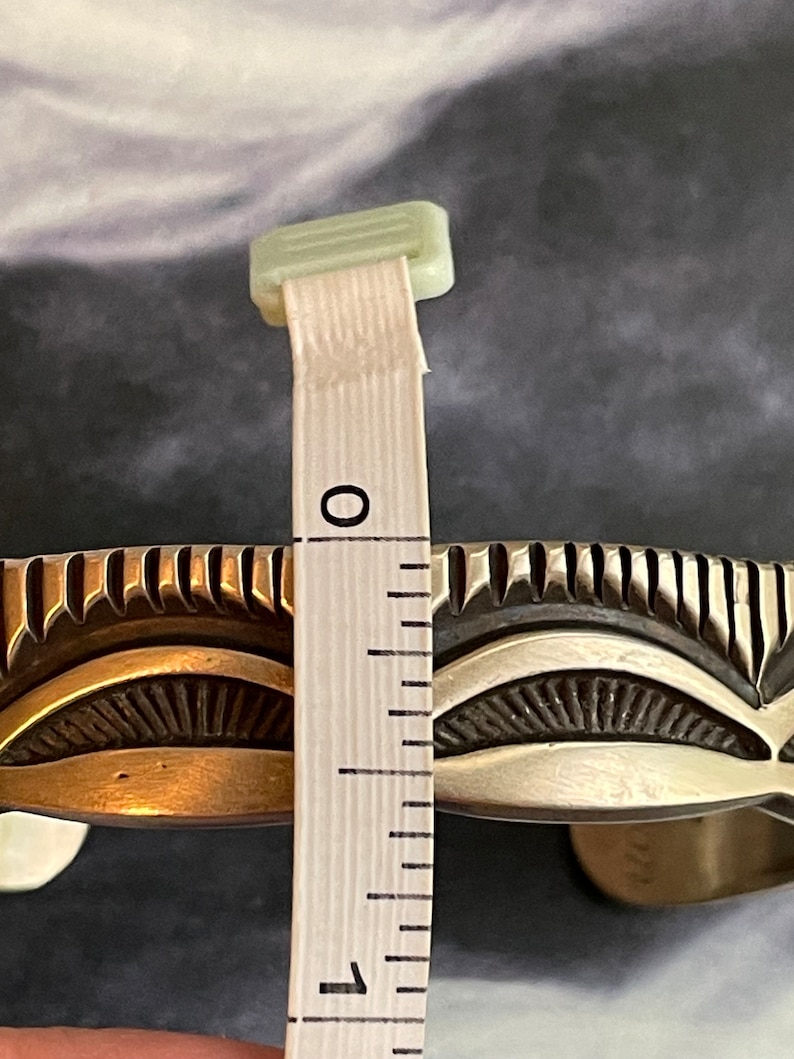 May include: A silver and brass Native American style cuff bracelet with a decorative design. The bracelet is measured with a ruler, showing it is approximately 1 inch wide.