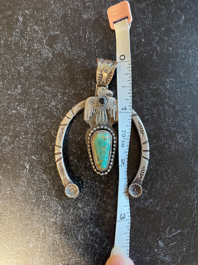 May include: A silver pendant featuring a turquoise stone and a bird motif. The pendant measures approximately 7.6 cm in length. It has a horseshoe shape with floral details at the ends.