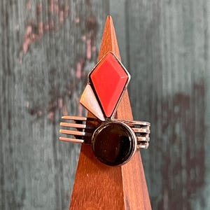 May include: A silver ring with a geometric design. The ring features a red diamond-shaped stone, a black circular stone, and a white triangular stone. The ring is displayed on a wooden stand against a green and brown background.