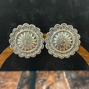 May include: A pair of round silver-toned earrings featuring a central sunburst design surrounded by a rope-like texture and a scalloped edge. The metal surface has a polished finish with darkened recessed areas to highlight the intricate details.