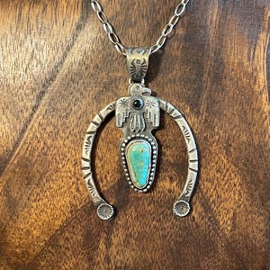 May include: Silver pendant necklace with a turquoise stone and a stylized bird design. The pendant has a horseshoe shape with decorative details and a black stone accent. The necklace hangs on a chain.