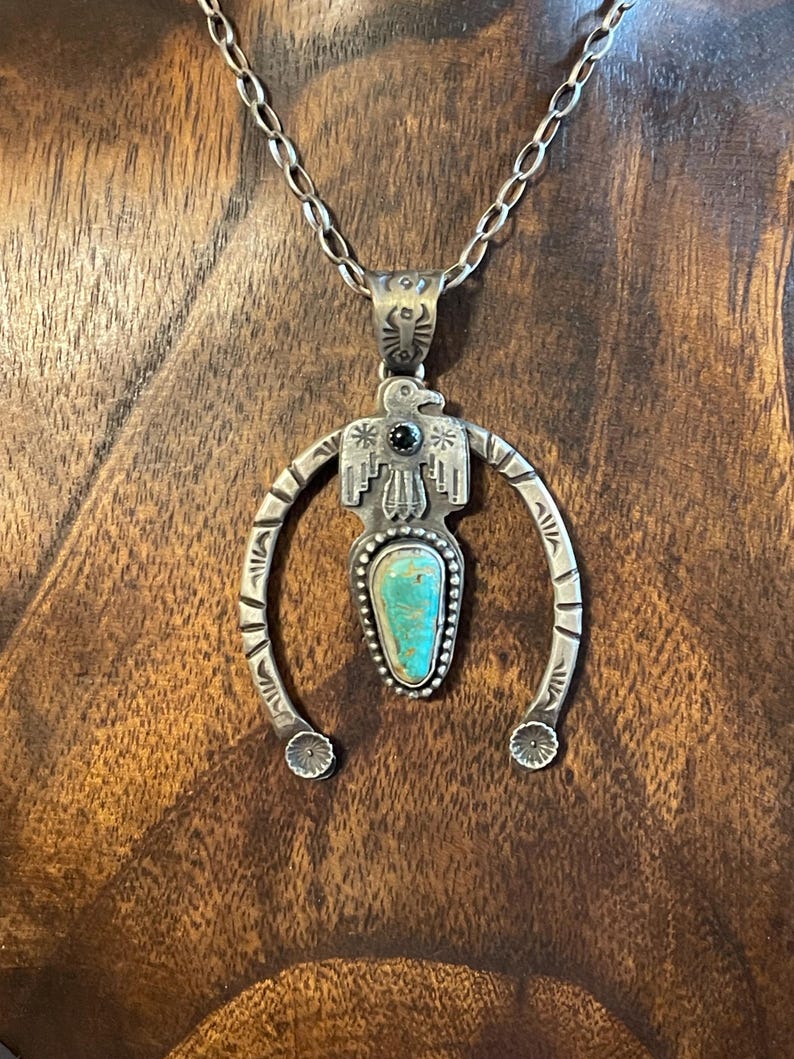 May include: Silver pendant necklace with a turquoise stone and a stylized bird design. The pendant has a horseshoe shape with decorative details and a black stone accent. The necklace hangs on a chain.