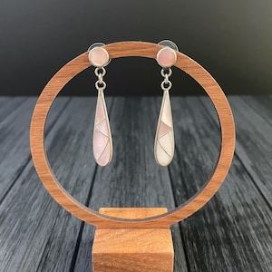 May include: A pair of silver earrings with a geometric design. Each earring features a round pink stud, a chain, and a teardrop-shaped pendant with pink and white segments. The earrings are displayed on a wooden stand against a dark background.