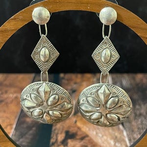 May include: A pair of silver-toned drop earrings featuring a circular stud connected to a diamond-shaped charm with etched lines and a central oval, hanging above a larger circular pendant embossed with a symmetrical floral-inspired pattern.