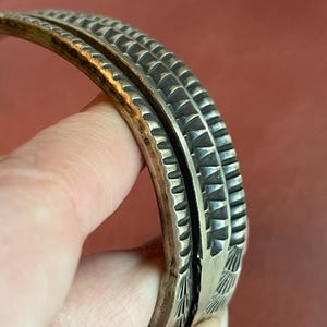 K&ouml;nnte beinhalten: Ein silbernes Armband mit komplizierten geometrischen Mustern. Das Armband besteht aus zwei B&auml;ndern, die miteinander verbunden sind. Die B&auml;nder sind mit einer Reihe von erhabenen Dreiecken und Linien verziert.