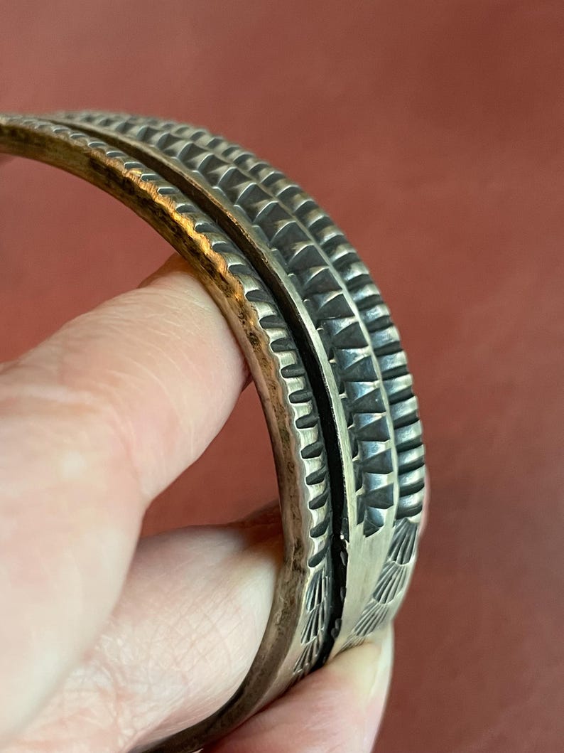 K&ouml;nnte beinhalten: Ein silbernes Armband mit komplizierten geometrischen Mustern. Das Armband besteht aus zwei B&auml;ndern, die miteinander verbunden sind. Die B&auml;nder sind mit einer Reihe von erhabenen Dreiecken und Linien verziert.