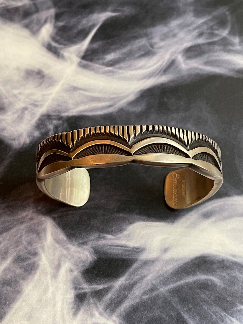 May include: A silver cuff bracelet with a geometric design. The bracelet is stamped with the word "Enoskin".
