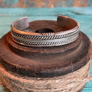 May include: A silver cuff bracelet with a detailed rope design and a central decorative element. The bracelet is displayed on a dark, textured surface, with a background of teal and brown tones. The cuff is approximately 2.5 inches wide.