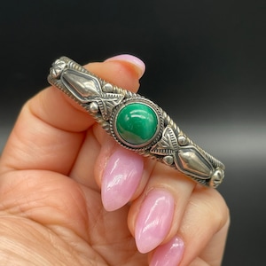 May include: A silver bracelet with a green gemstone set in the center. The bracelet has a detailed design with intricate carvings.