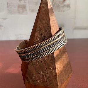 K&ouml;nnte beinhalten: Ein breites, silberfarbenes Armreif mit einem strukturierten, geometrischen Design. Das Armband wird auf einem Holzst&auml;nder pr&auml;sentiert.