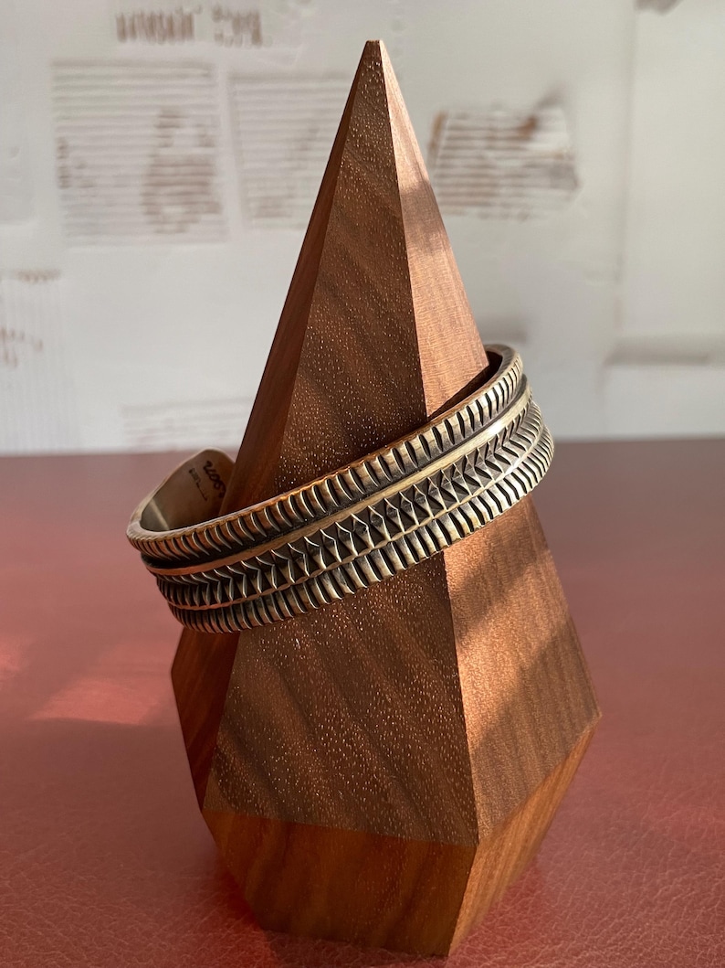 K&ouml;nnte beinhalten: Ein breites, silberfarbenes Armreif mit einem strukturierten, geometrischen Design. Das Armband wird auf einem Holzst&auml;nder pr&auml;sentiert.