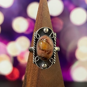 May include: A silver ring with an oval, multi-colored stone. The stone has shades of orange, brown, and pink. The ring has a decorative silver setting with a rope-like design around the stone and small silver accents. The ring is displayed on a wooden stand.