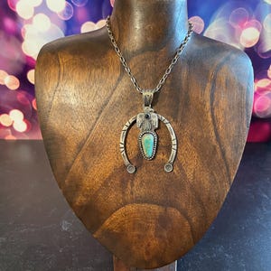 May include: A silver necklace with a turquoise pendant, presented on a wooden bust. The pendant has a central turquoise stone and a decorative frame. The necklace hangs from a delicate chain. The bust is set against a blurred background.