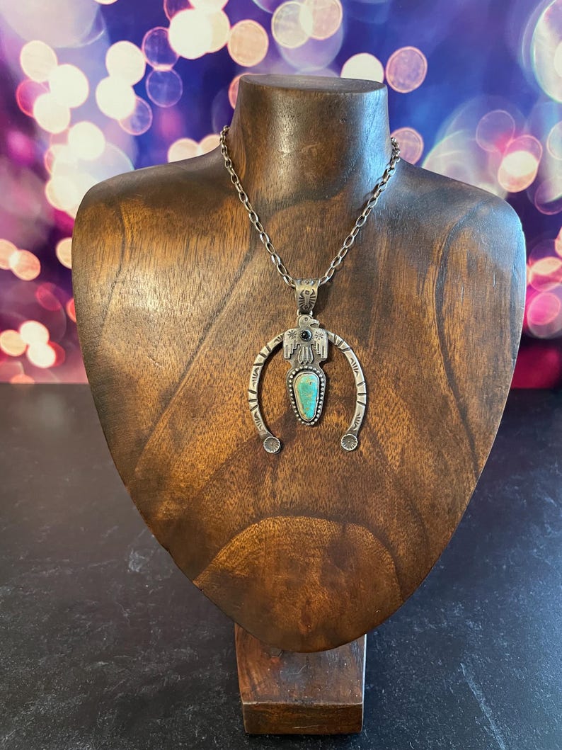May include: A silver necklace with a turquoise pendant, presented on a wooden bust. The pendant has a central turquoise stone and a decorative frame. The necklace hangs from a delicate chain. The bust is set against a blurred background.