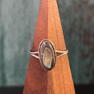 May include: A silver ring featuring an oval-shaped abalone shell inlay with iridescent green, blue, and brown tones. The center stone is framed by a twisted rope-style metal border and sits on a thin, split-shank silver band.