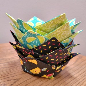 May include: A stack of handmade fabric bowl cozies in various patterns. The patterns include lime slices on turquoise, green swirls, and black with taco and confetti designs. The cozies are stacked on a wooden surface.
