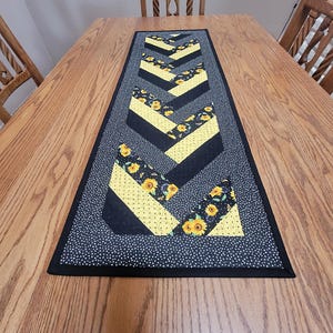 May include: Black and yellow chevron table runner with a sunflower print. The runner has a black border and white polka dots on the background fabric.