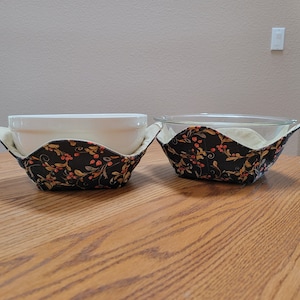 May include: Two fabric bowl cozies with a black background and a floral pattern in shades of orange, yellow, and white. The cozies are holding a white bowl and a clear glass bowl.
