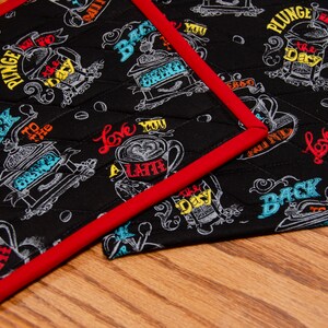 May include: Two black fabric pot holders with red trim. The pot holders have a white and colorful design with coffee-themed text and illustrations. The text includes "Back to the Grind", "Love You a Latte", and "Plunge into the Day".