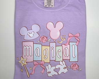 May include: A lavender Comfort Colors t-shirt featuring the word "magical" in pastel colors. The design includes Mickey Mouse-inspired shapes, a wand, bows, roses, and a balloon. The shirt is folded neatly on a white surface.