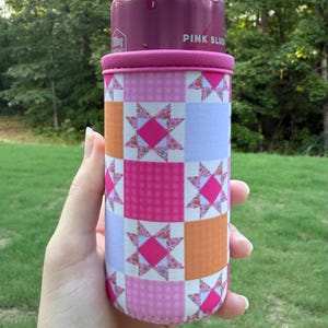 Puede incluir: Un enfriador de latas cil&iacute;ndrico de espuma con un patr&oacute;n de patchwork de cuadrados alternos en naranja s&oacute;lido, morado claro s&oacute;lido y cuadros vichy rosas. El dise&ntilde;o incluye motivos de estrellas en rosa y blanco. Contiene una lata de bebida rosa.