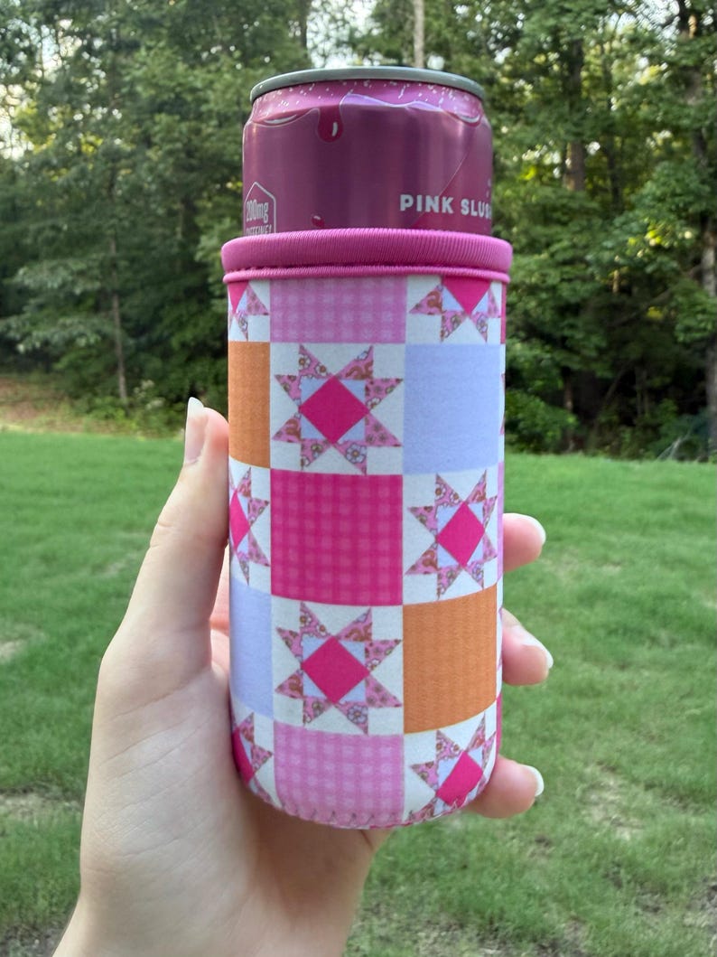 Puede incluir: Un enfriador de latas cil&iacute;ndrico de espuma con un patr&oacute;n de patchwork de cuadrados alternos en naranja s&oacute;lido, morado claro s&oacute;lido y cuadros vichy rosas. El dise&ntilde;o incluye motivos de estrellas en rosa y blanco. Contiene una lata de bebida rosa.