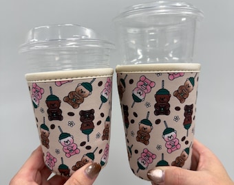 May include: Two clear plastic cups with lids are held in beige drink sleeves. The sleeves feature a repeating pattern of brown and pink teddy bears holding iced coffee drinks. The sleeves have a cream-colored trim.