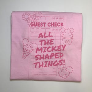 May include: A light pink t-shirt with a graphic design. The design includes the words "GUEST CHECK" and "ALL THE MICKEY SHAPED THINGS!" in pink, along with illustrations of Mickey Mouse-shaped treats.