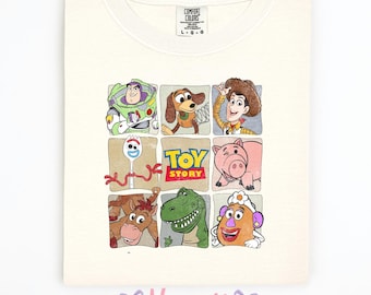 May include: Cream-colored Comfort Colors t-shirt featuring a grid of Toy Story characters including Buzz Lightyear, Woody, and others. The Toy Story logo is centered. The shirt has a crew neck and short sleeves.