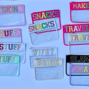 May include: Assortment of clear plastic zippered pouches with colorful glitter lettering. The pouches have various phrases including "SKIN", "SNACKS", "TRAVEL", "STUFF", "WIFEY BRIDE", "GLAM", and "MAKEUP".