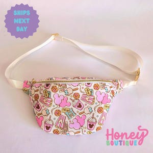May include: A white fanny pack with a pink adjustable strap. The fanny pack has a zipper closure and is covered in a print of Mickey Mouse ears, popcorn buckets, ice cream cones, and other Disney-themed snacks.